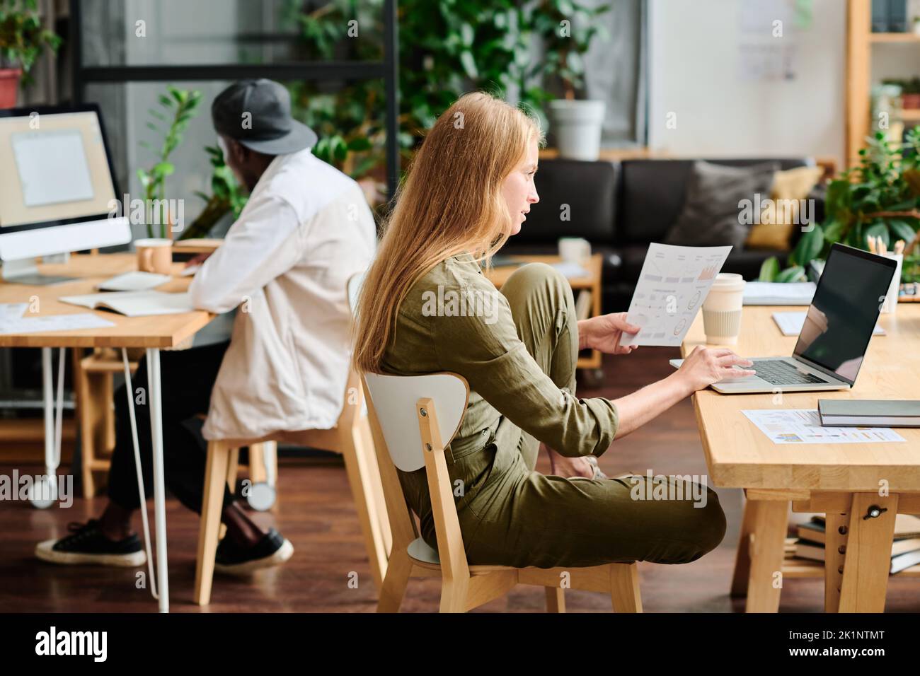 Joven empresaria con documento financiero mirando la pantalla del portátil mientras se sienta junto al lugar de trabajo y analizando la información en línea Foto de stock