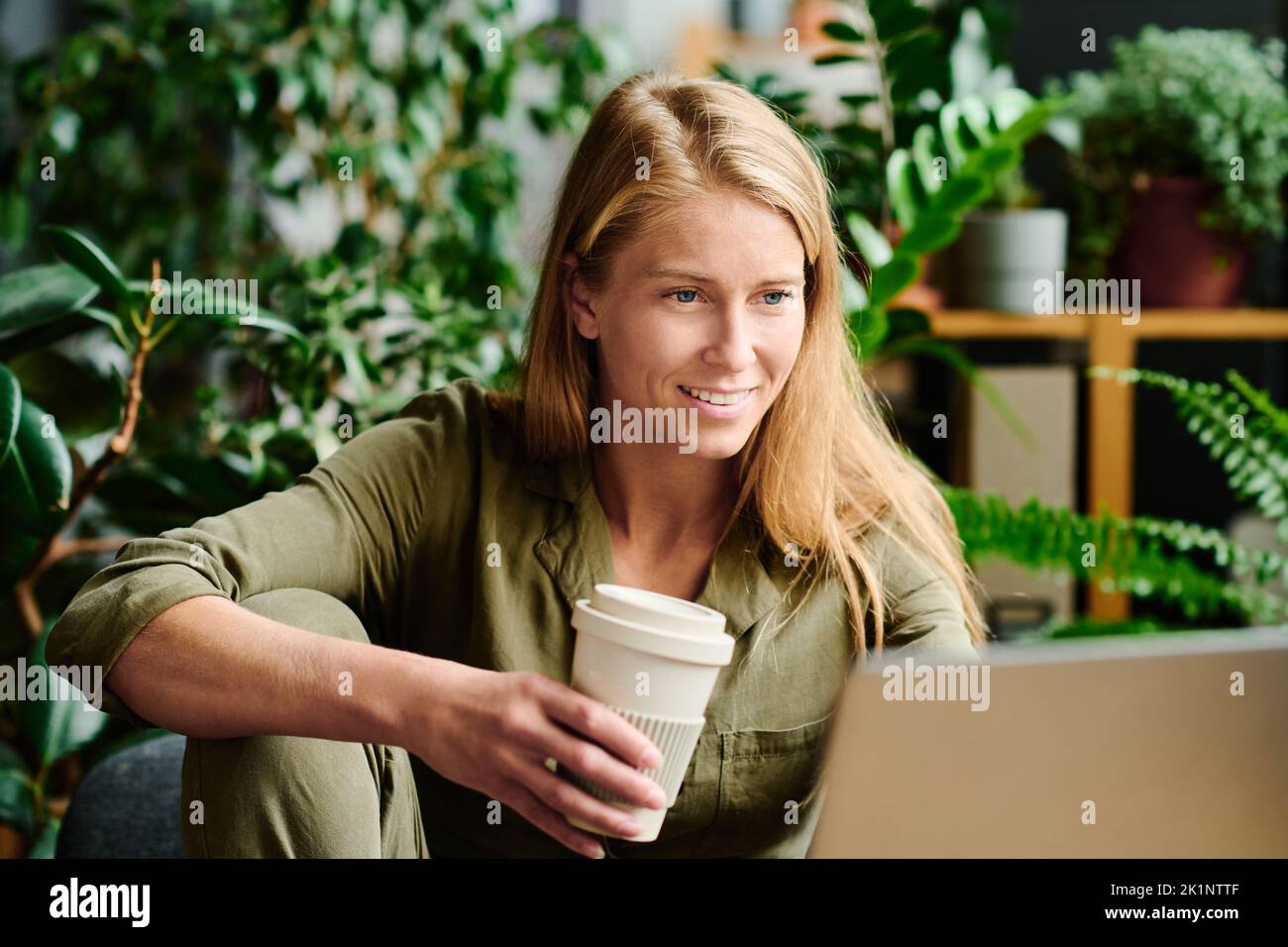 Joven mujer de negocios sonriente mirando la pantalla del ordenador portátil mientras se comunica en chat de vídeo y tomando café en el lugar de trabajo Foto de stock