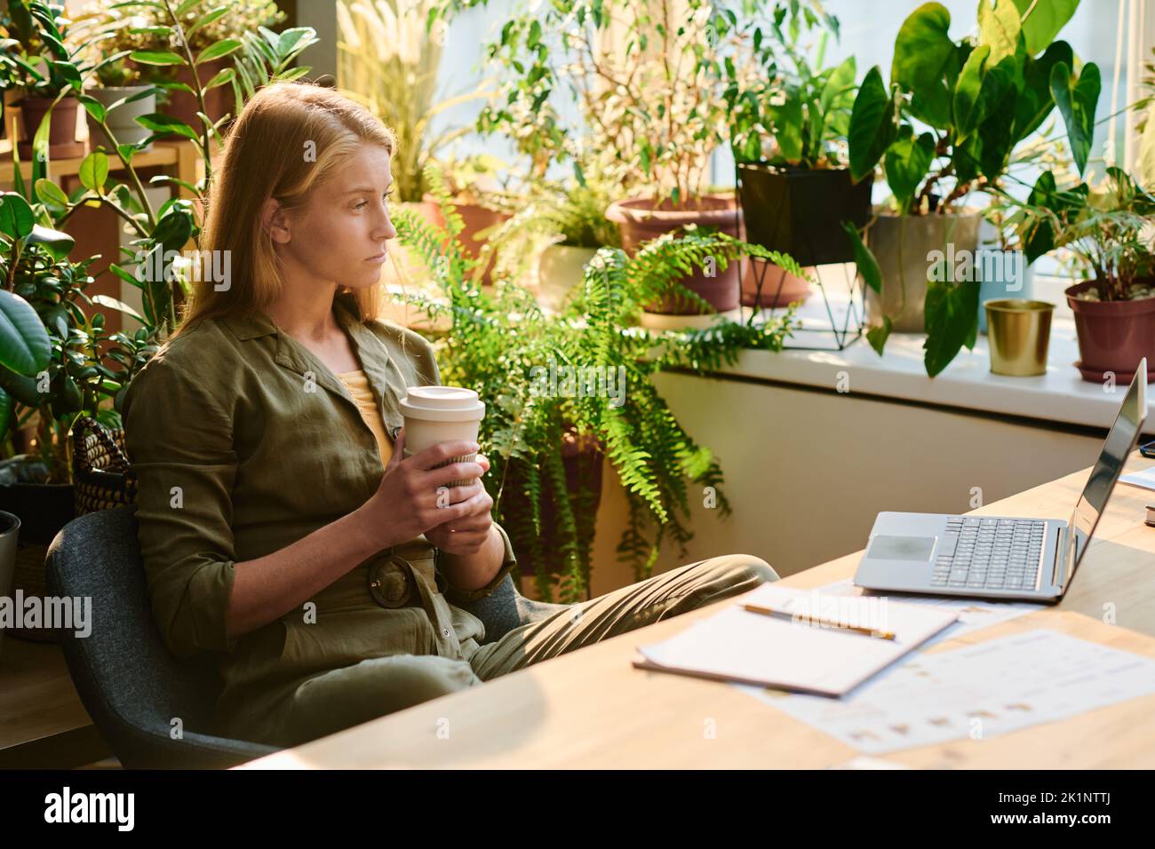 Mujer joven rubia con una taza de café mirando la pantalla del portátil durante la conexión en red o mientras ve un vídeo en línea o se comunica Foto de stock