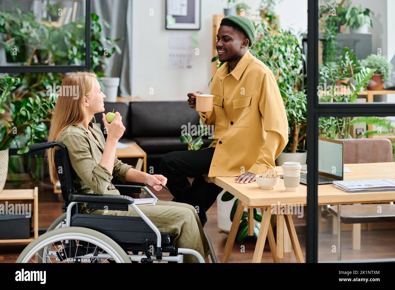 Dos empleados jóvenes de interculturalidad discutiendo los puntos de trabajo durante el almuerzo mientras toman aperitivos y café o té en la oficina verde Foto de stock