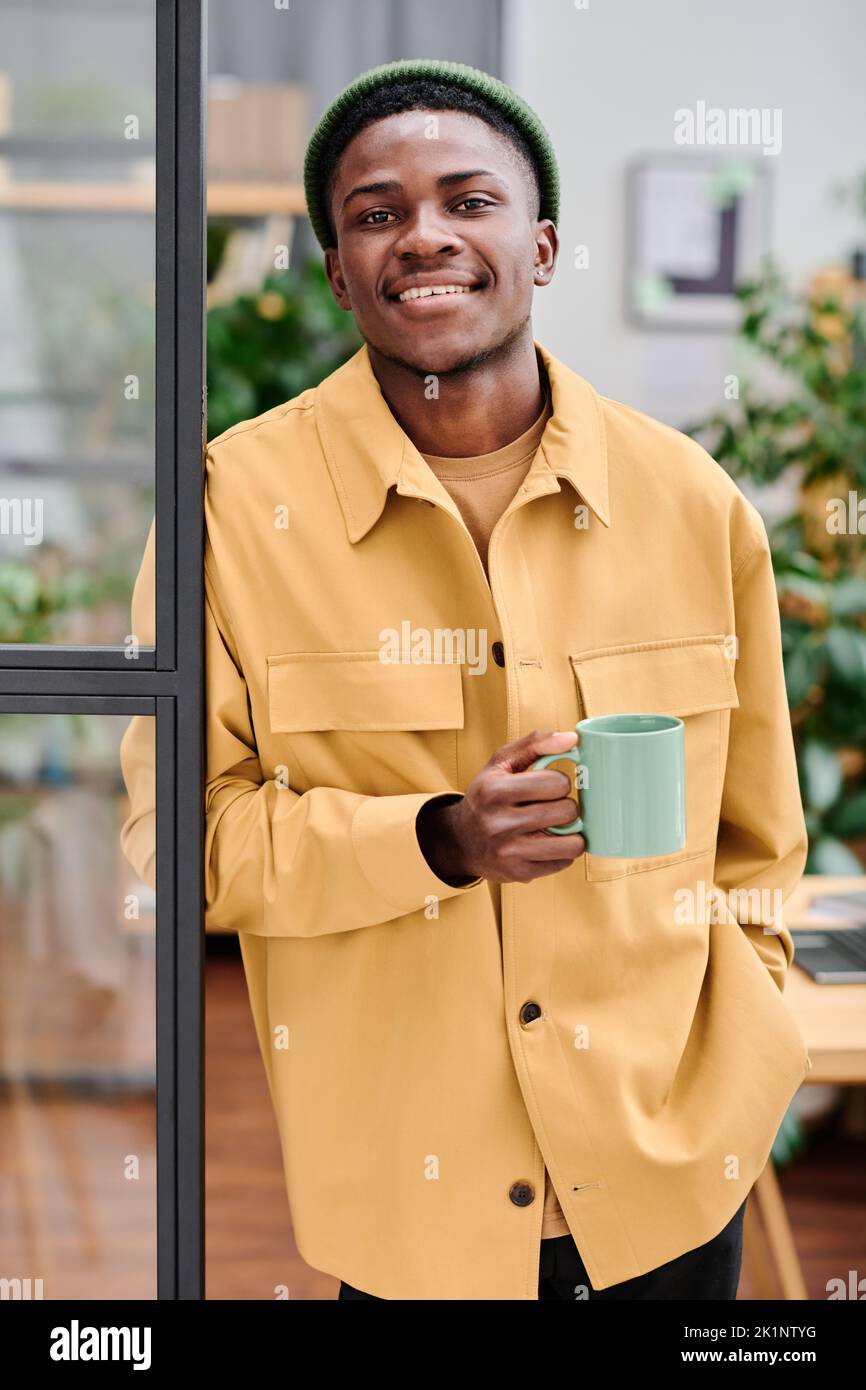 Joven exitoso hombre de negocios afroamericano con una taza de café delante de la cámara contra la oficina del espacio abierto Foto de stock