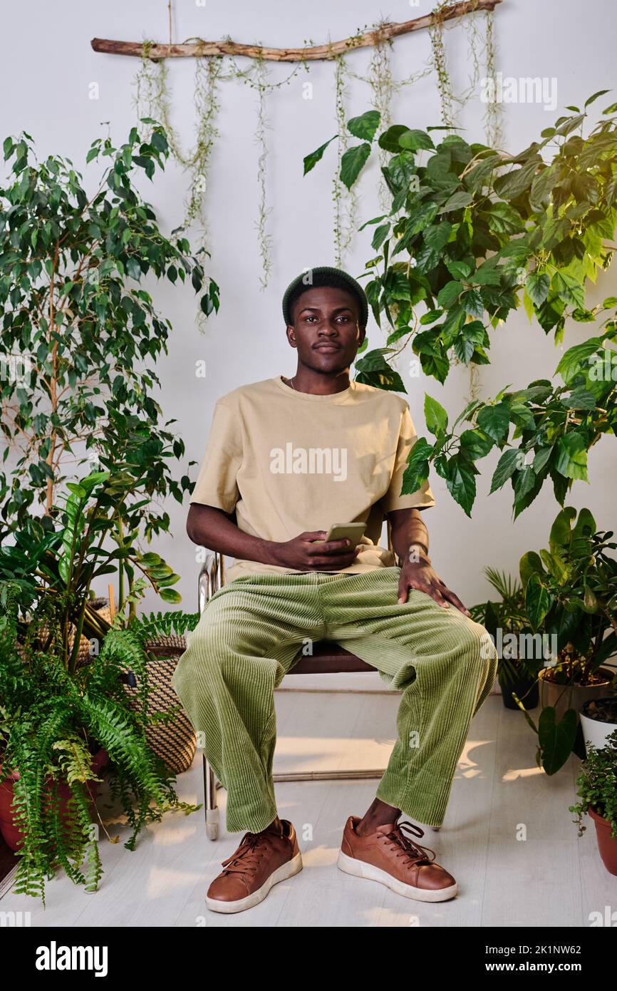 Hombre afroamericano joven en ropa casual con smartphone sentado en silla entre plantas verdes en la oficina y mirando la cámara Foto de stock