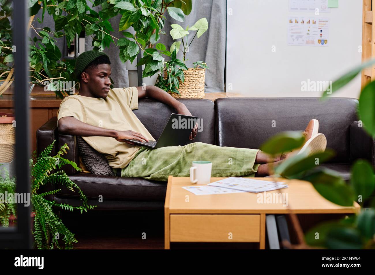 Joven hombre relajado en ropa casual con el ordenador portátil en las rodillas sentado en un sofá de cuero negro y buscando a través de los datos en línea para un nuevo proyecto Foto de stock