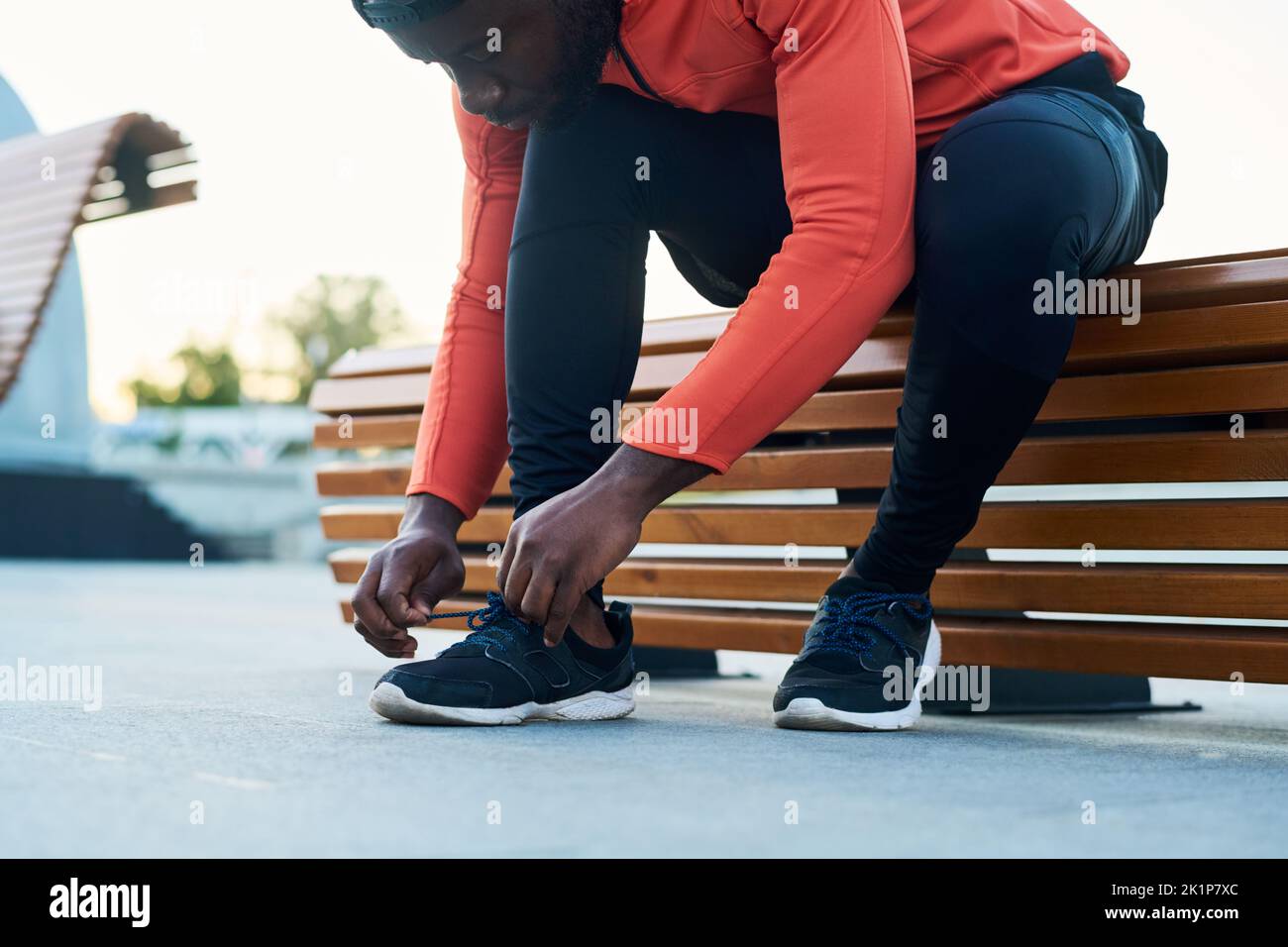 Atleta joven o corredor con chaqueta deportiva roja y leggins negras atando el zapato de las sneakers mientras se sienta en un banco de madera Foto de stock