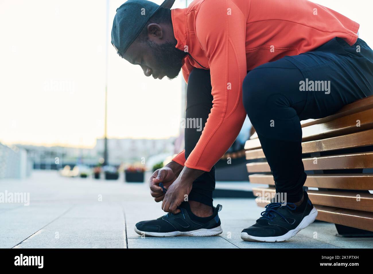 Vista lateral del joven corredor afroamericano con leggins y gorra negras y chaqueta deportiva roja que se dobla sobre las sneakers mientras ata el zapato Foto de stock