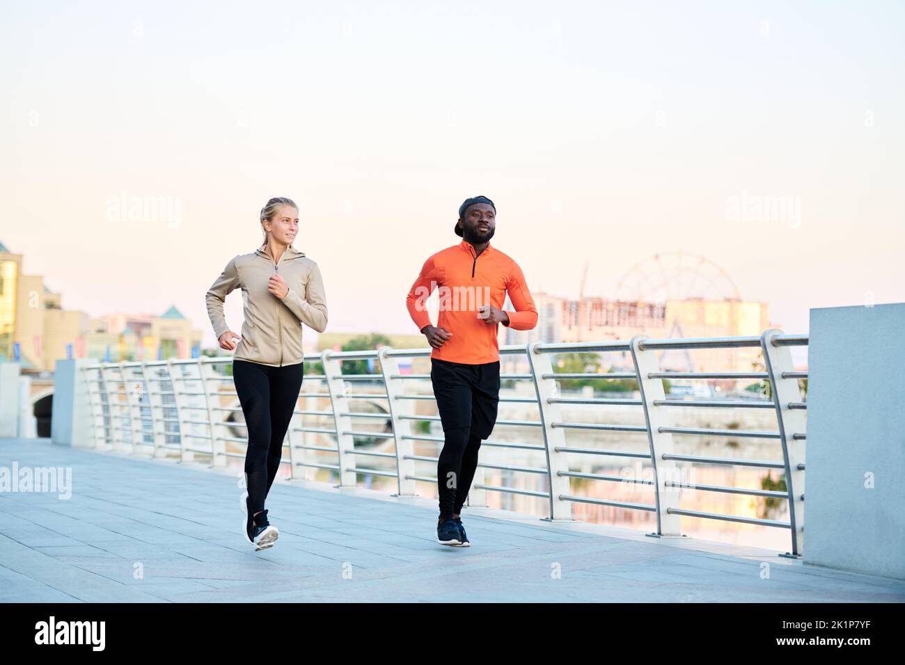 Dos jóvenes atletas interculturales en chaquetas deportivas, leggins y zapatillas de deporte para correr a lo largo del puente junto al río por la mañana Foto de stock