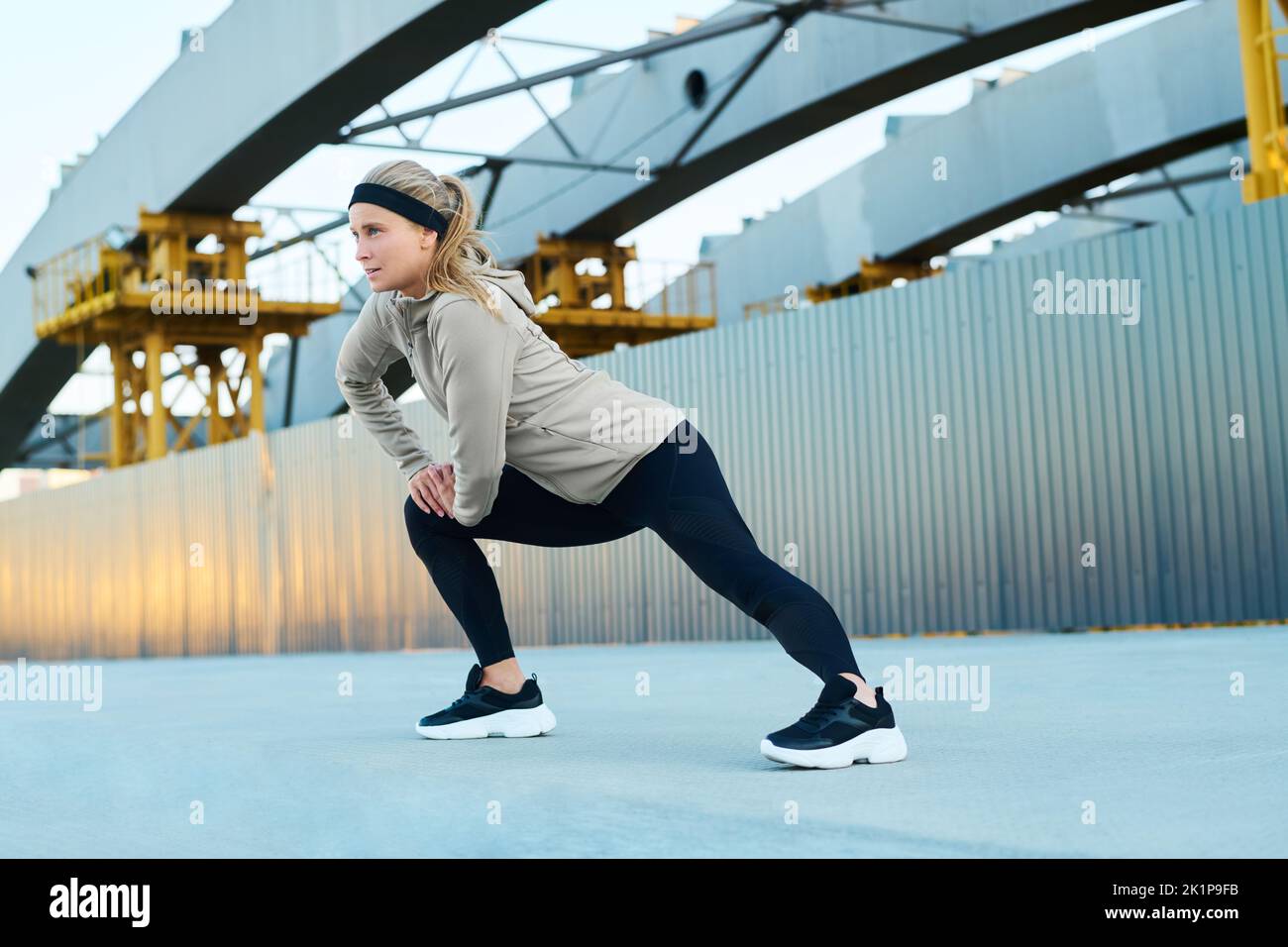 Joven mujer rubia en ropa activa haciendo ejercicio físico para estirar la pierna mientras está de pie sobre asfalto contra la arquitectura moderna Foto de stock