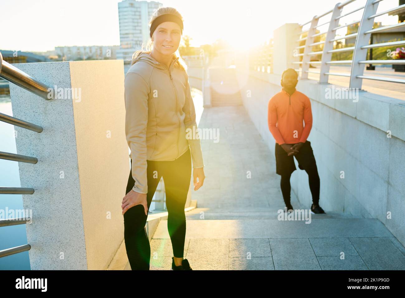 Feliz joven deportista rubia en ropa activa parada en el puente junto a la orilla del río contra la luz del sol y atleta afroamericano Foto de stock