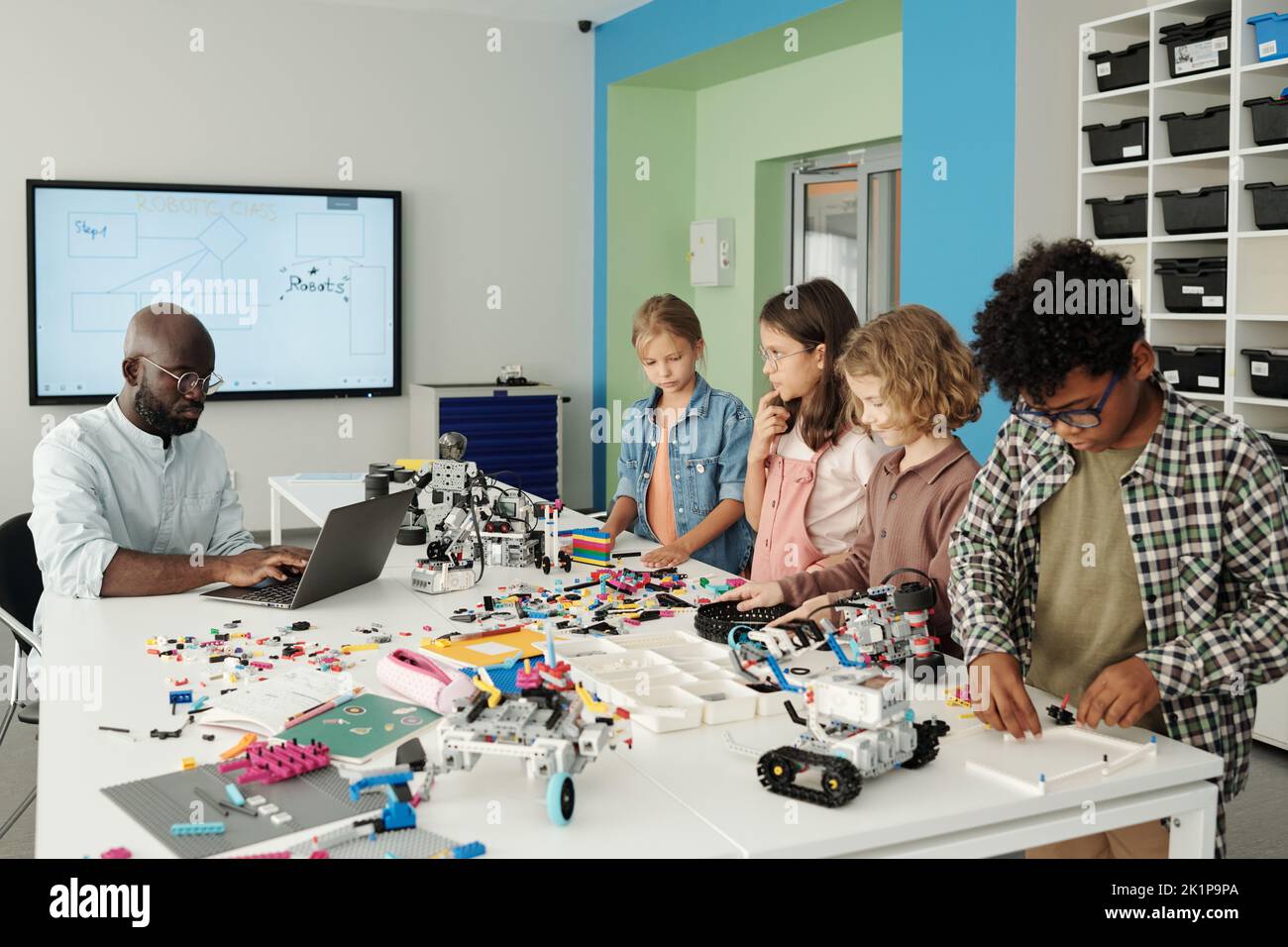 Grupo de escolares multiculturales delante de un maestro afroamericano de robótica escribiendo en el teclado del ordenador portátil durante la lección Foto de stock