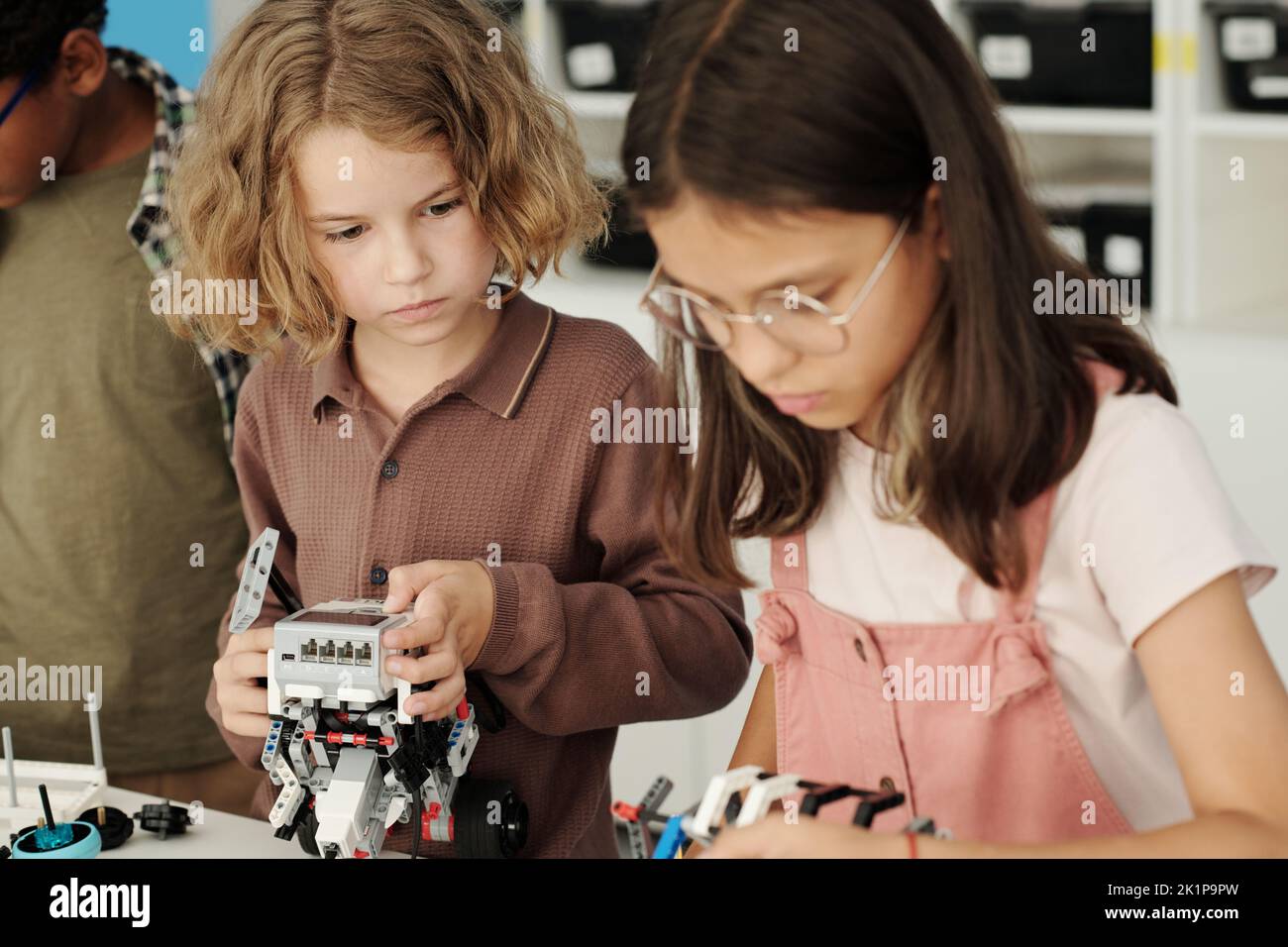 Coqueta colegiala mirando a su compañero de clase construyendo el robot mientras ambos están de pie junto a la mesa con detalles para la conexión en la lección Foto de stock