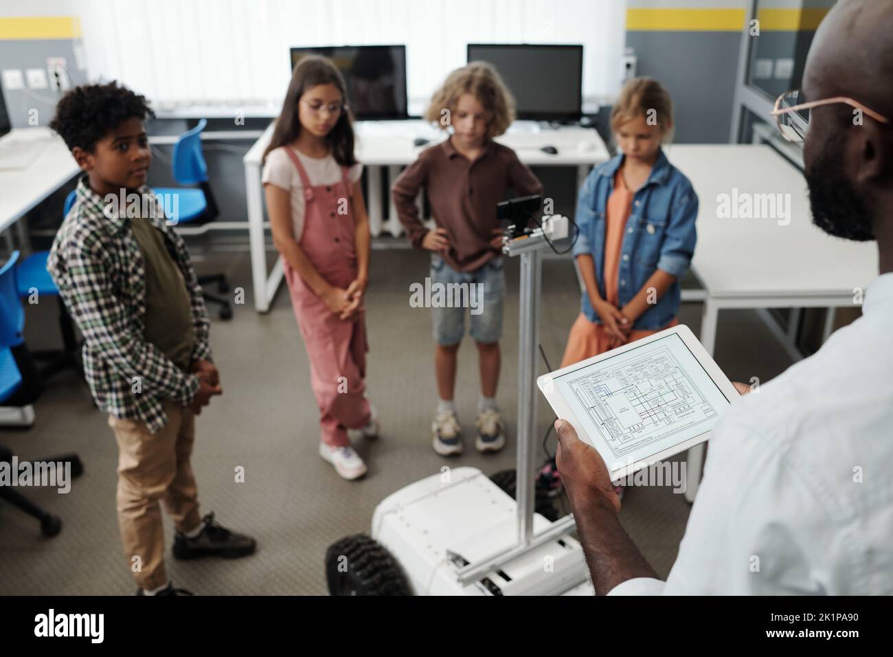 Profesor afroamericano con la tableta de pie frente a los alumnos y robot rastreador mientras hace la presentación del nuevo juguete en la lección Foto de stock