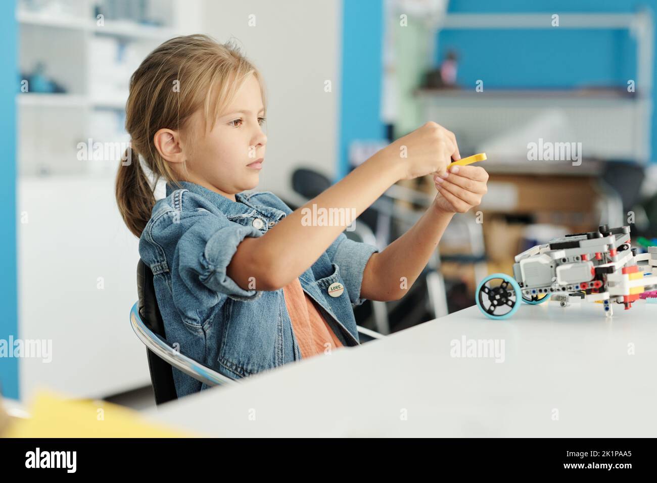 Bonita niña en ropa casual que conecta los detalles de juguete electrónico mientras se sienta en el escritorio en la lección de robótica y la creación de un nuevo robot Foto de stock
