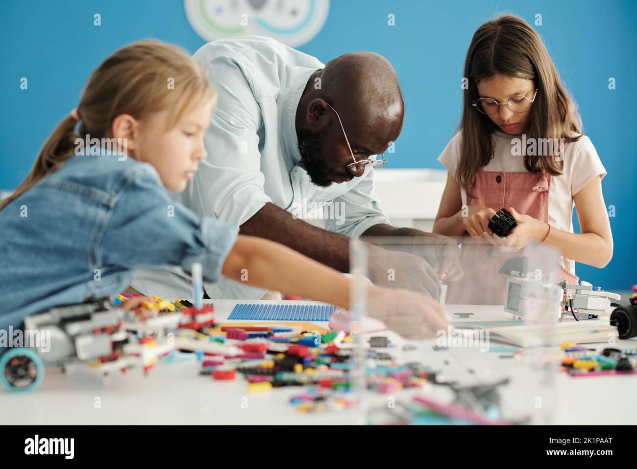 Joven afroamericano inclinándose sobre el escritorio con los detalles mientras ayuda a los estudiantes de primaria a conectar partes de robots Foto de stock
