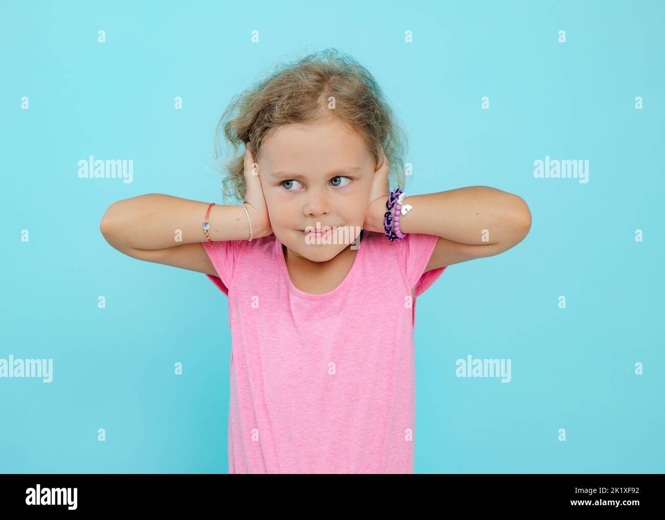 Pequeña pequeña expresiva, conmocionada, descontenta rubia rubia rizada en camisa rosa mirar lejos y cubrir las orejas con las manos en el estudio Foto de stock