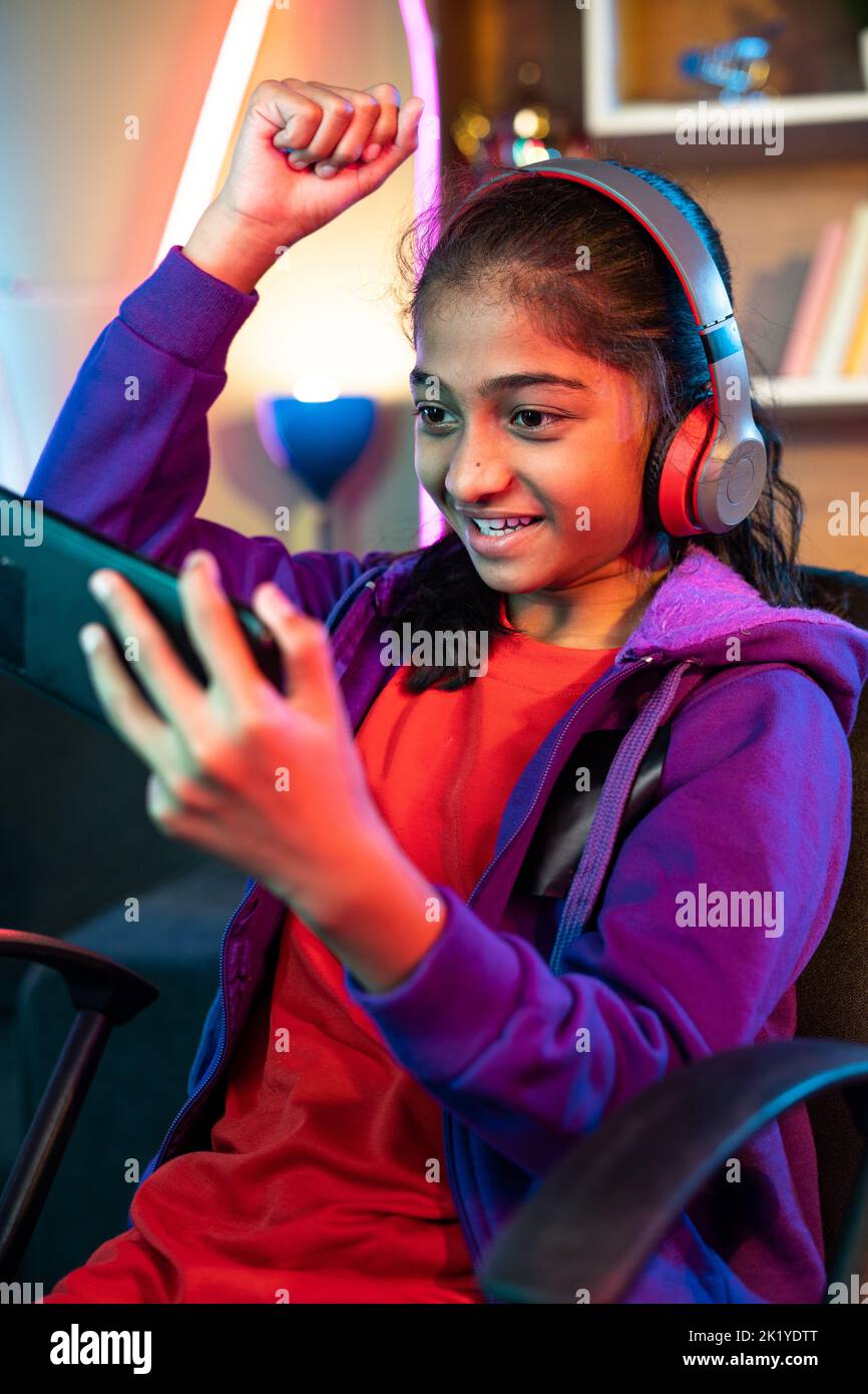Fotografía vertical de una joven muy emocionada con auriculares que celebran la victoria mientras juega a videojuegos en el teléfono móvil en casa: Concepto de logros Foto de stock