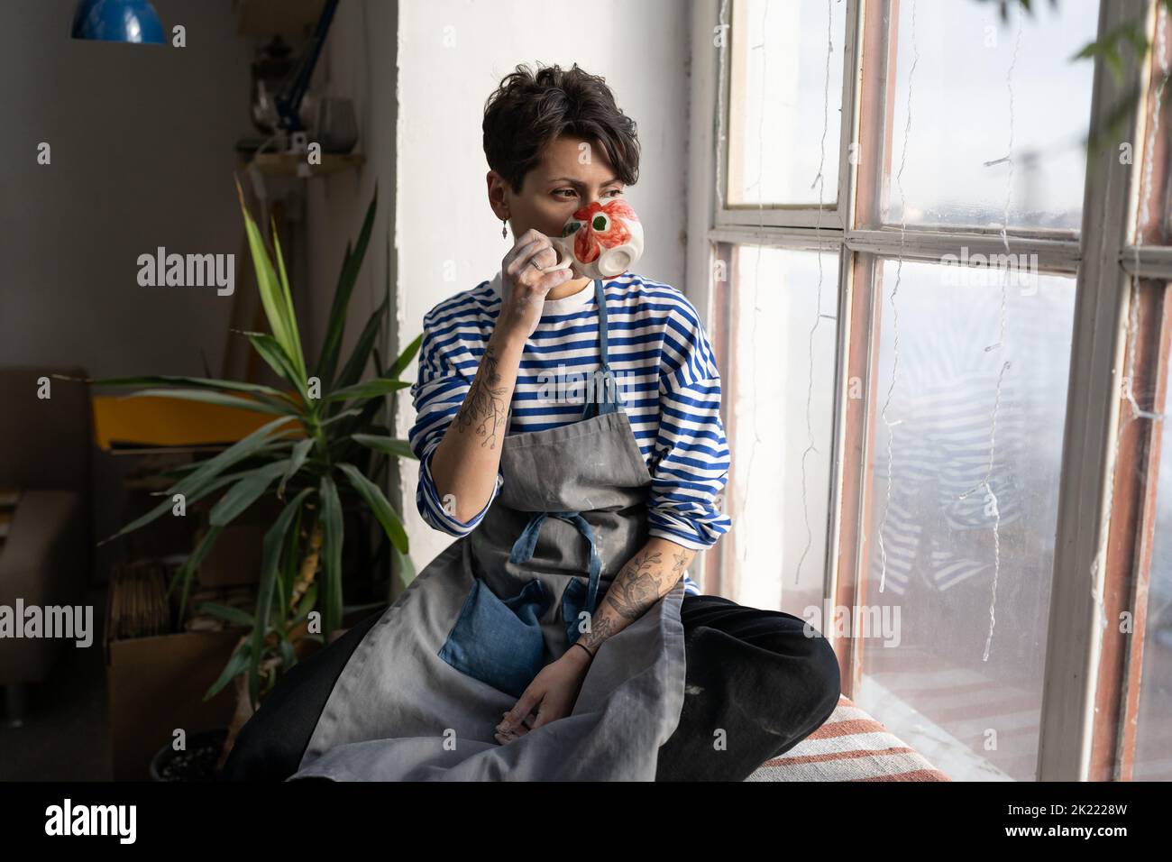 Alfarero hembra en delantal sucio sosteniendo una taza de arcilla hecha a mano con café, disfrutando de un descanso en el taller Foto de stock
