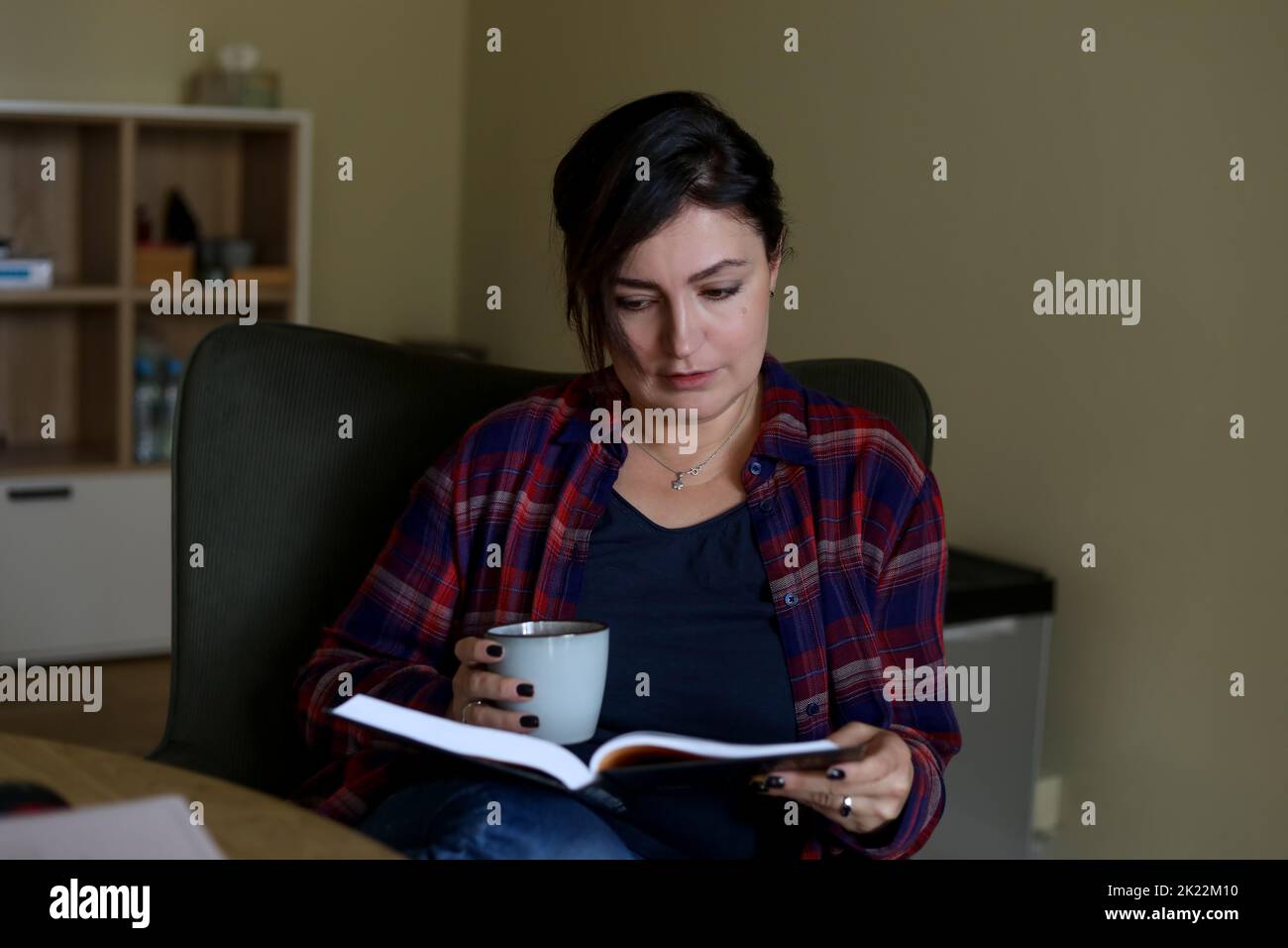 Mujer morena Relájese con un libro y una taza de té en el sillón del armario de la oficina. Actividad de lectura para mujeres adultas, disfrutando de un estilo de vida tranquilo en el interior Foto de stock