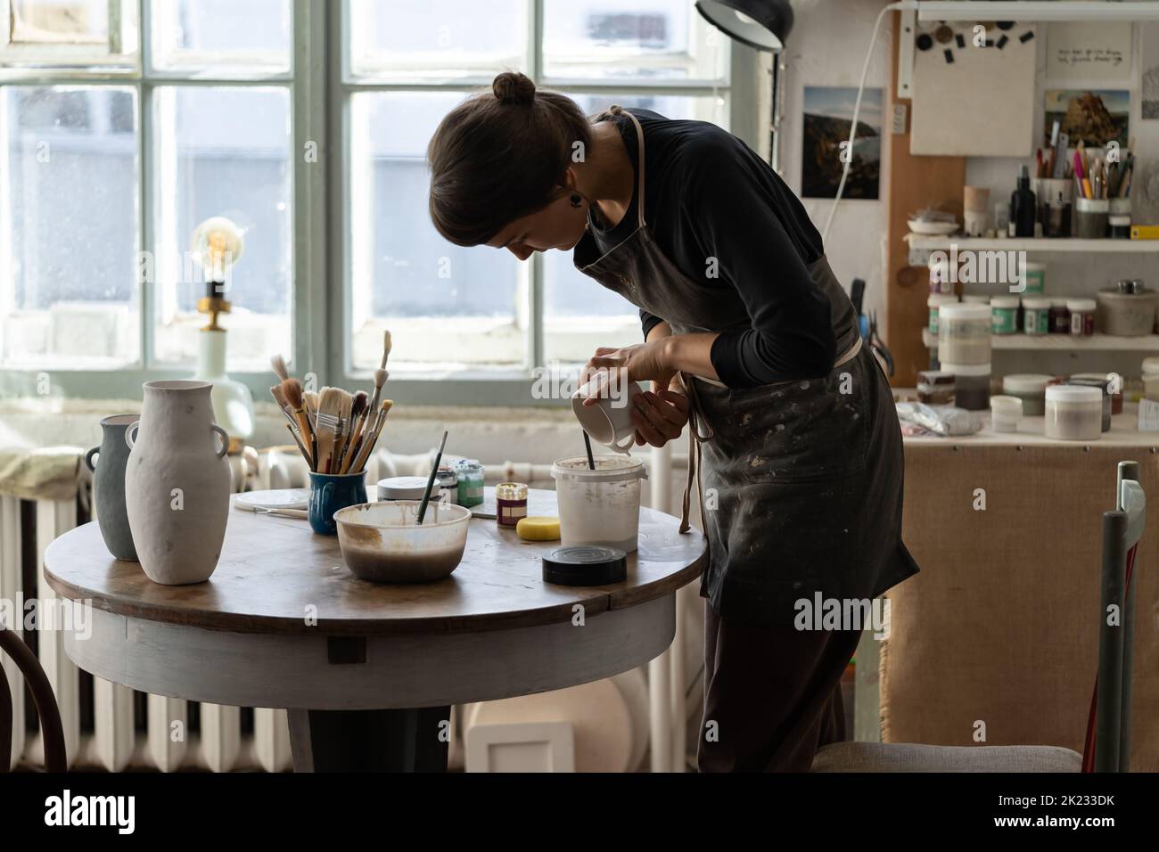 Mujer morena en delantal negro vierte pintura blanca en taza de cerámica para trabajar con la cerámica en el taller Foto de stock