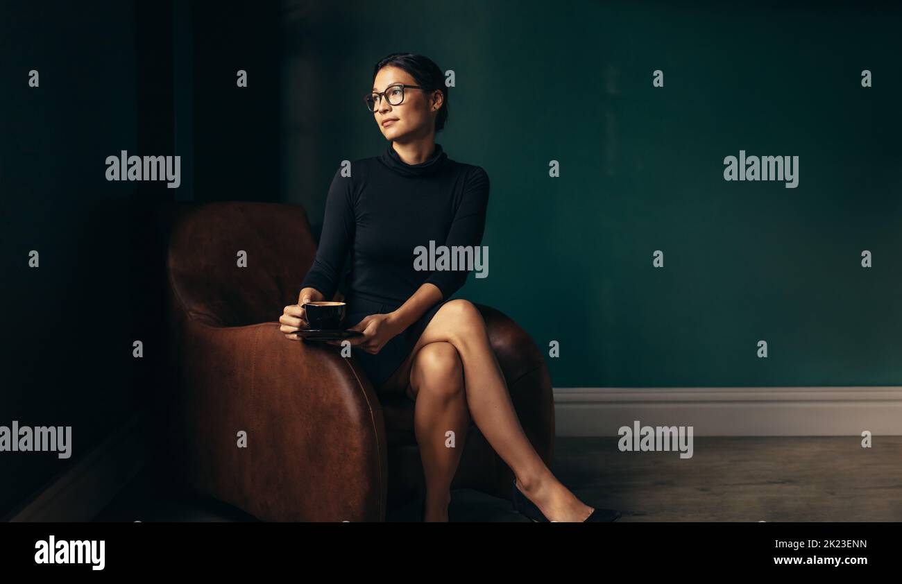 Mujer asiática sentada en un sillón con una taza de café y mirando a otro lado. Pensativa mujer de negocios relajándose en su oficina. Foto de stock