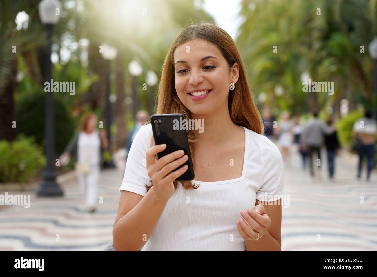 Mujer joven emocionada viendo su smartphone mientras camina por la calle con personas borrosas en el fondo. Niña milenaria con aplicación móvil al aire libre. Concepto de tecnología de estilo de vida para adolescentes. Foto de stock