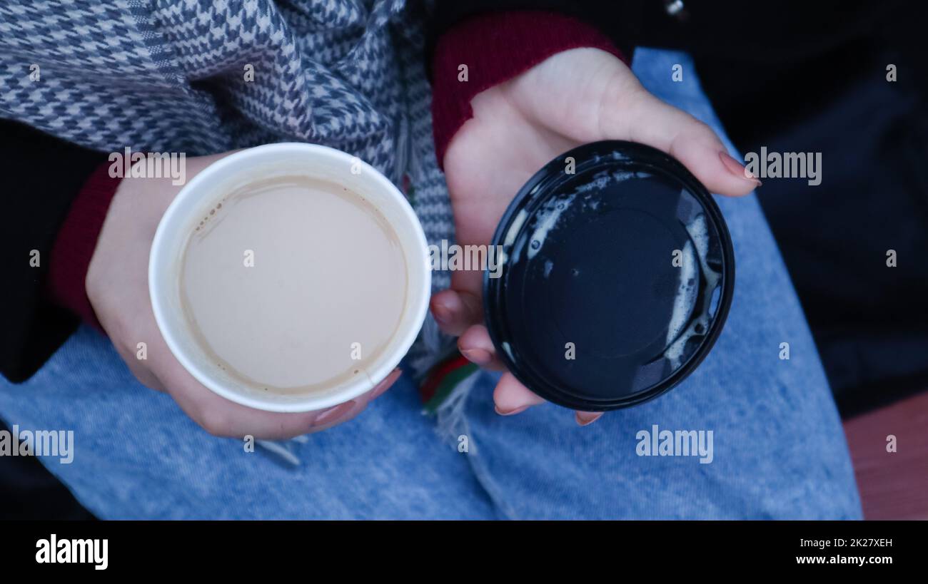 Las manos femeninas están sosteniendo el café de la mañana para llevar y cerrando una taza de papel con una tapa mientras se sienta en un banco del parque, cerca de las manos. Foto de stock