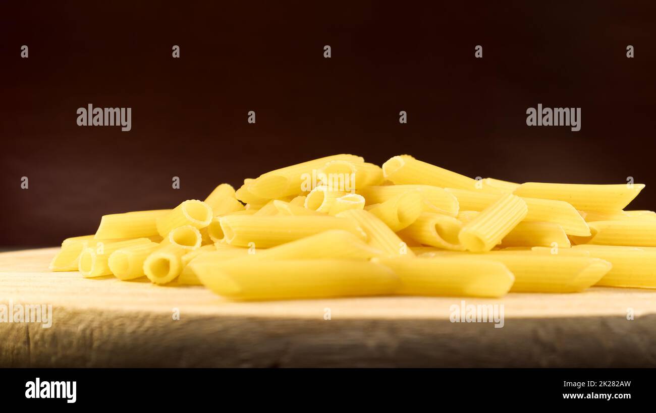 La pasta cruda Penne Rigate es una pasta corta con cortes oblicuos y una superficie estriada. Pasta tradicional italiana. Pasta de fondo. Vista lateral de un ingrediente italiano en un plato de madera. Foto de stock