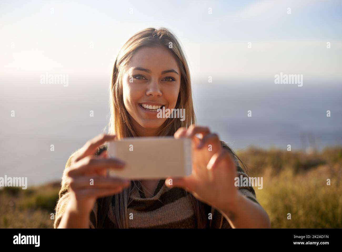 Un momento que vale la pena recordar. Retrato de una joven atractiva sosteniendo su teléfono móvil para hacer una foto al aire libre. Foto de stock