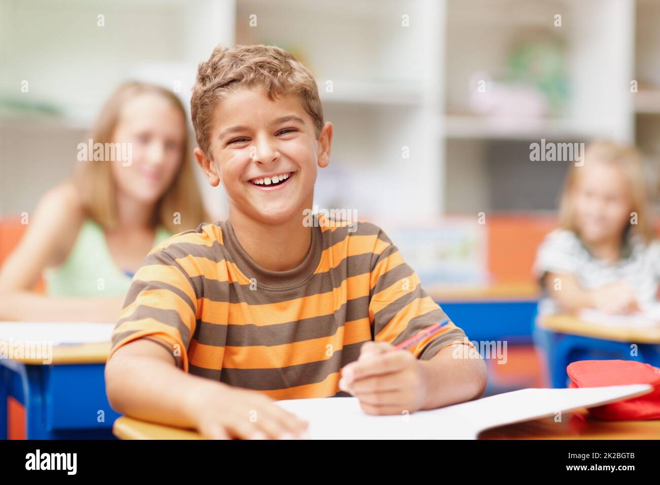 Disfrutar de un momento ligero en clase. Dulce chico sentado en su escritorio en clase riendo felizmente. Foto de stock