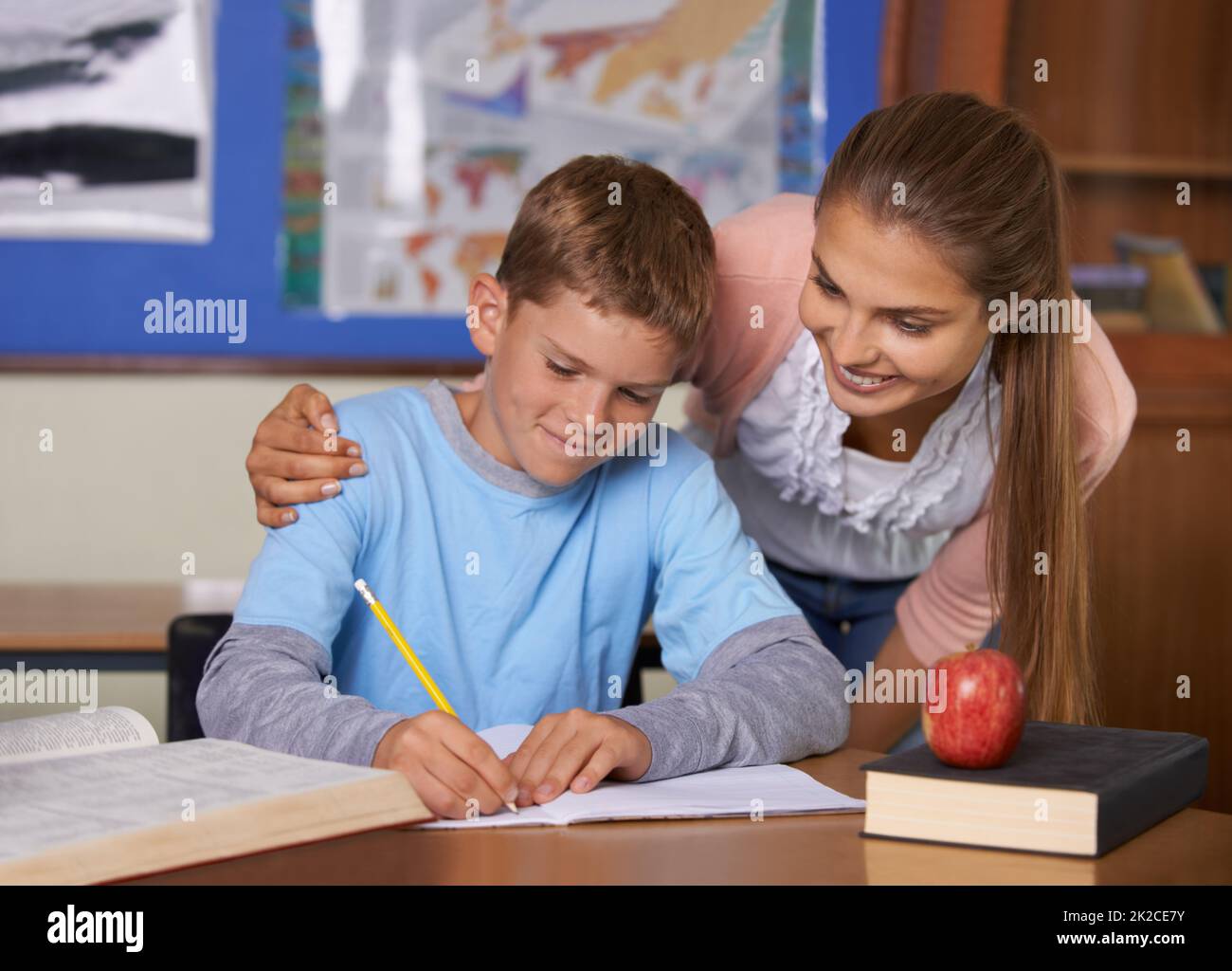 Cada niño es especial. Una joven maestra de apoyo que se inclina sobre su estudiante mientras escribe en un libro. Foto de stock