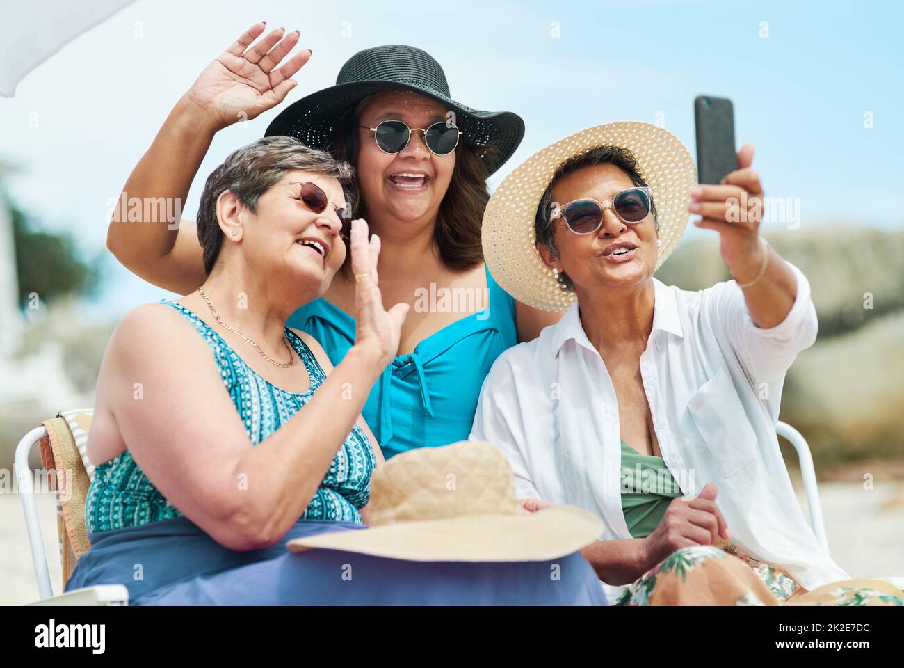 Mire cómo buena mamá se ve en un traje de baño. Foto de un grupo maduro de amigos usando un teléfono móvil para un chat de vídeo durante un día en la playa. Foto de stock