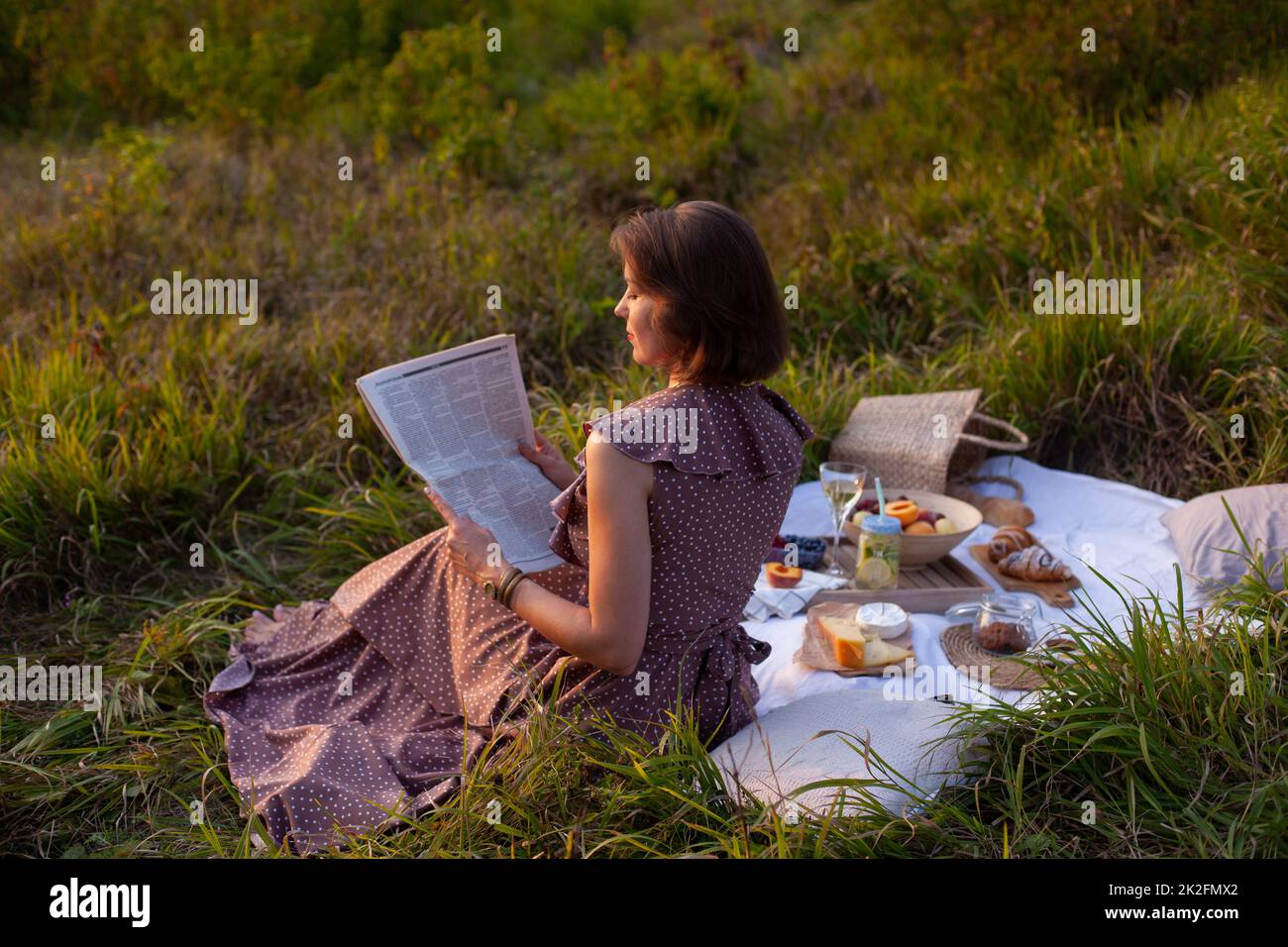 Una mujer con un largo vestido de verano con pelo corto sentado sobre una manta blanca y leyendo el periódico. Concepto de hacer un picnic en un parque de la ciudad durante su Foto de stock