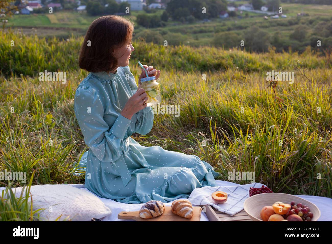 Una mujer con un largo vestido de verano con pelo corto sentado sobre una manta blanca con frutas y pasteles y limonada para beber. Concepto de hacer un picnic Foto de stock