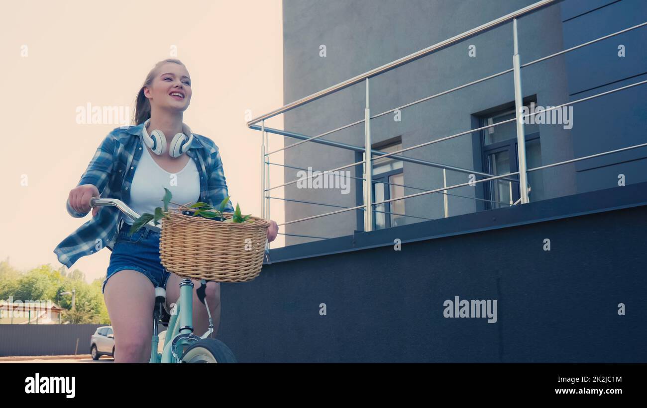 Ángulo bajo de visión de mujer joven feliz con auriculares en bicicleta en la calle, imagen de archivo Foto de stock