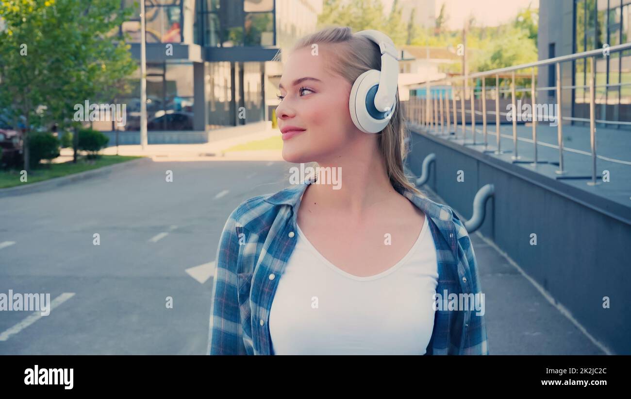 Mujer positiva en camisa de cuadros escuchando música con auriculares en la calle urbana, imagen de stock Foto de stock