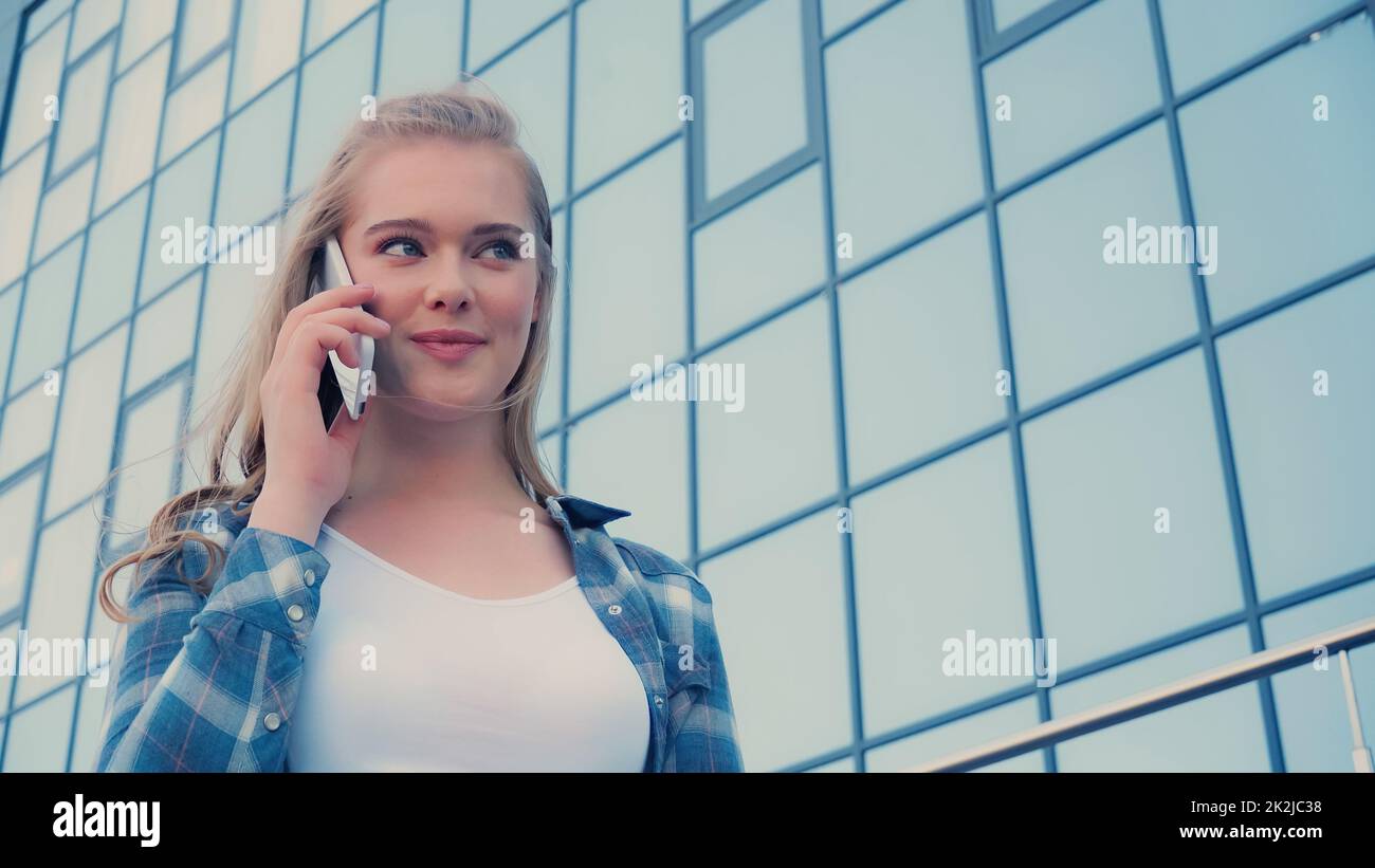 Ángulo bajo de visión de una mujer rubia sonriente hablando en el exterior del smartphone, imagen de archivo Foto de stock