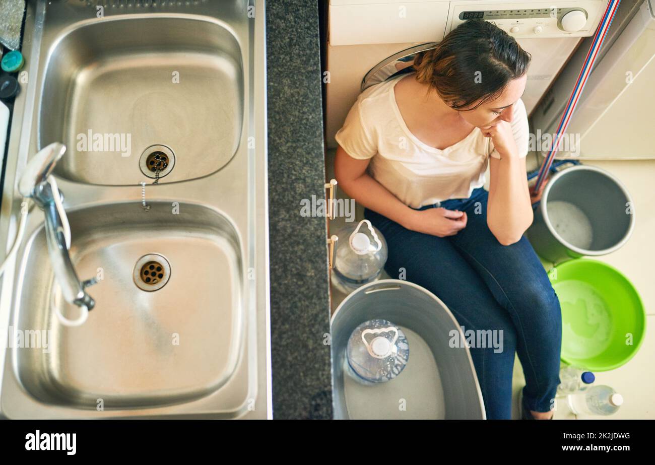 Preocupado por el agua. Imagen en ángulo alto de una hermosa mujer joven que se siente deprimida por la crisis del agua mientras se sienta en el suelo de la cocina en casa. Foto de stock