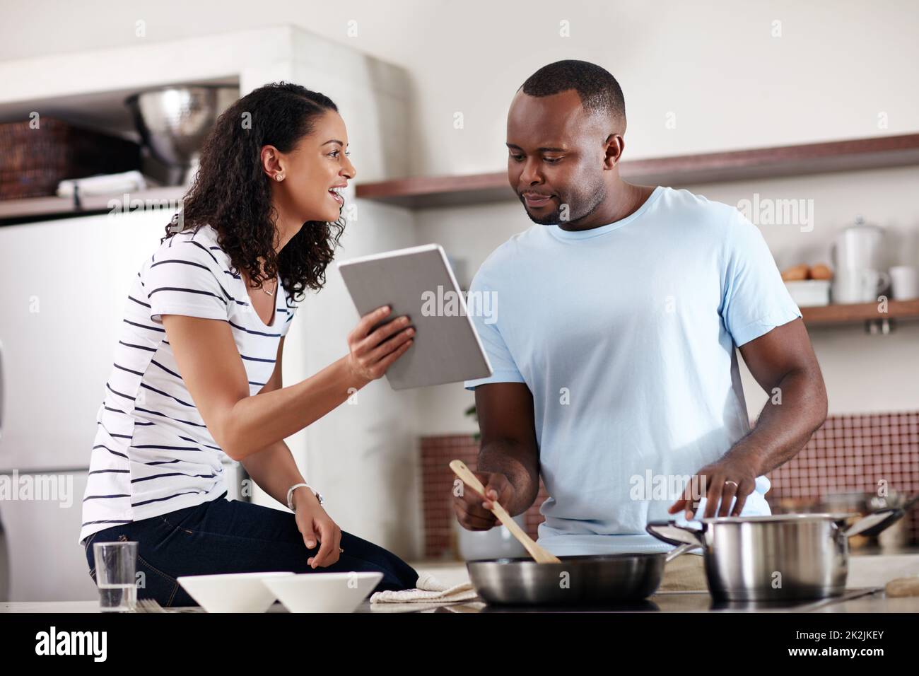 Veamos lo que dice la aplicación de cocina. Recortó la foto de una joven pareja casada usando una tableta mientras cocinaba juntos en la cocina de casa. Foto de stock