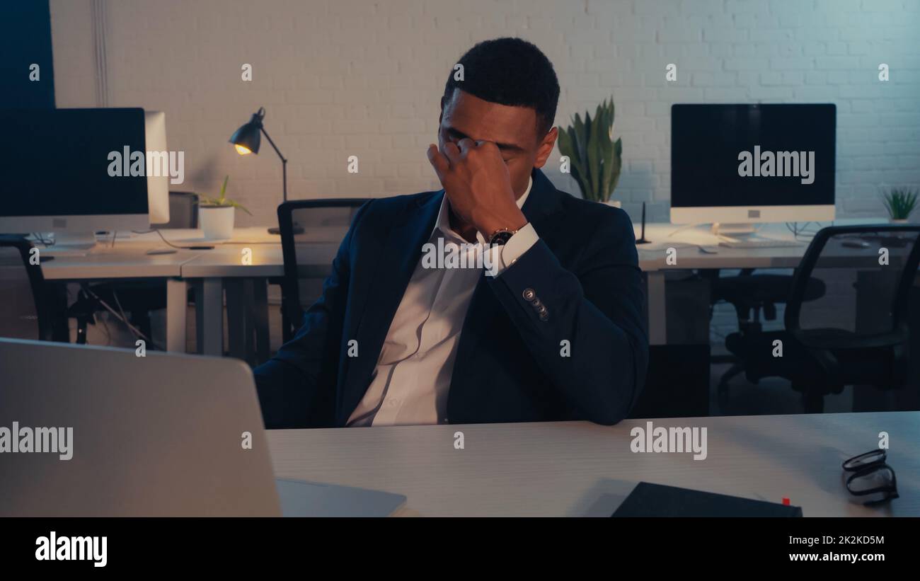 Exhausto hombre de negocios afroamericano sentado cerca del ordenador portátil y gafas en la oficina por la noche, imagen de archivo Foto de stock