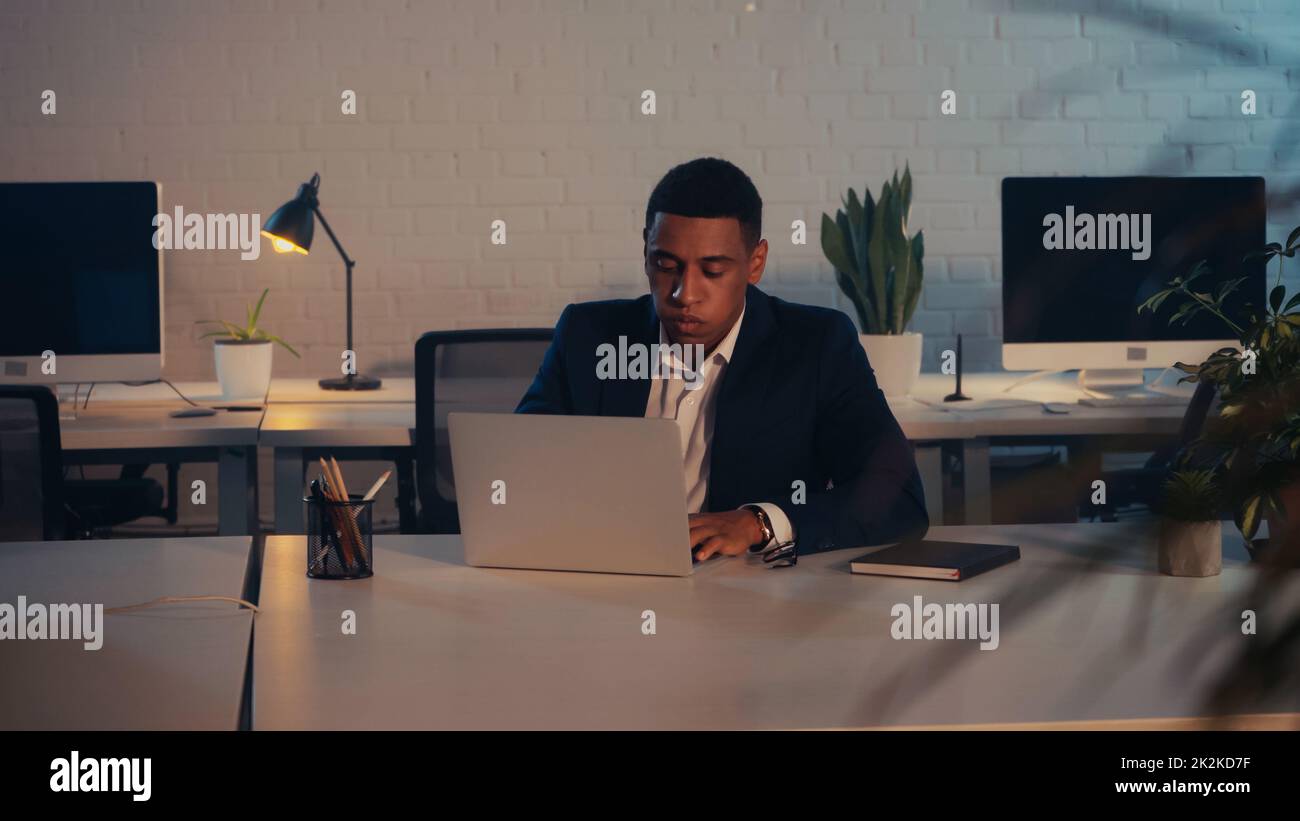 exhausto hombre de negocios afroamericano usando el portátil en la oficina por la noche, imagen de archivo Foto de stock