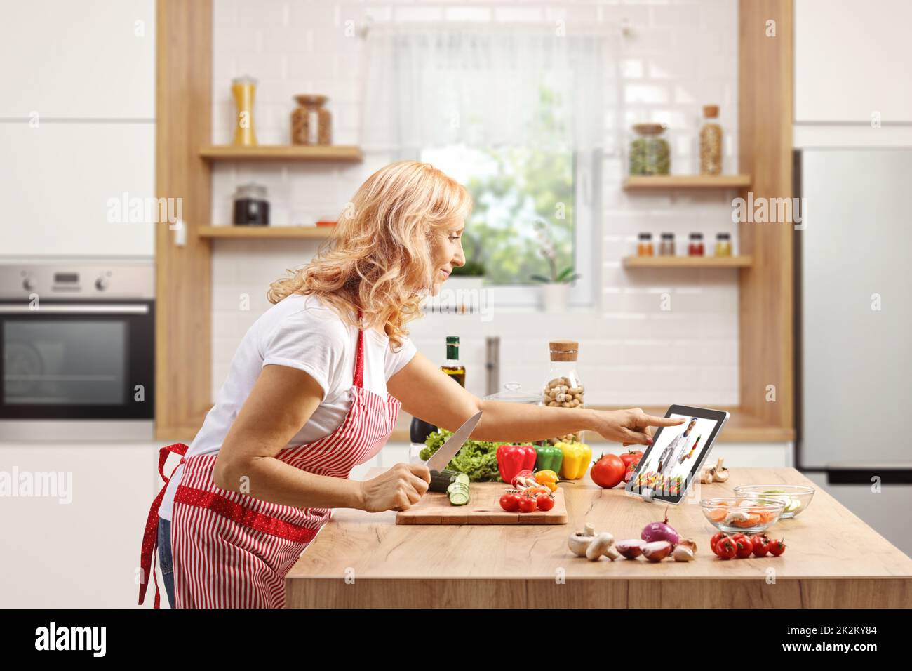 Mujer madura cocinando en un mostrador y viendo un vídeo en una tableta digital dentro de una cocina Foto de stock