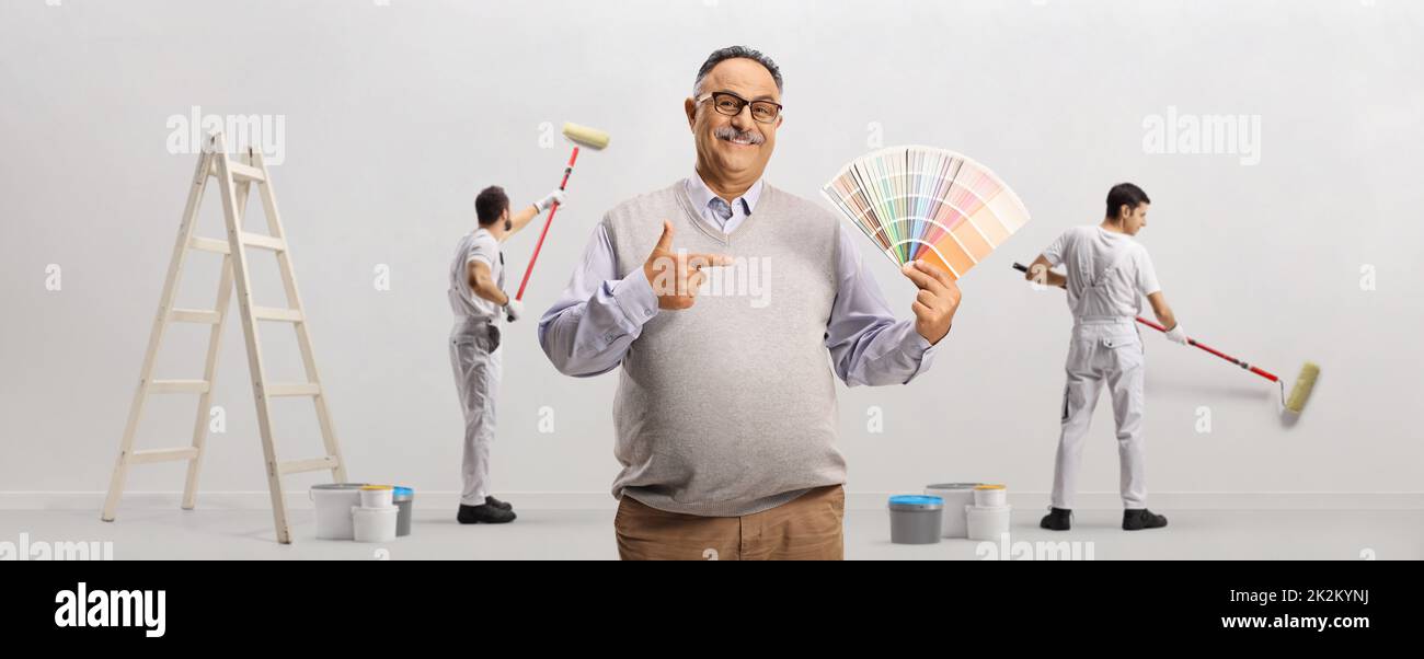 Hombre maduro sosteniendo una paleta de colores y pintores pintando una pared en el fondo Foto de stock