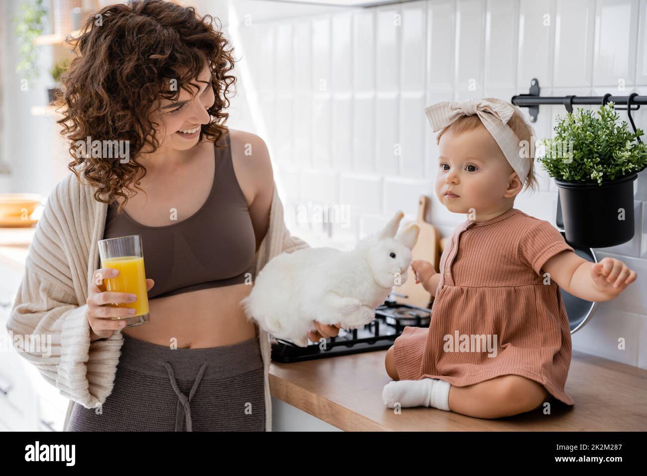niña sentada en la encimera de la cocina cerca de madre alegre con un vaso de jugo de naranja y un juguete suave, imagen de archivo Foto de stock