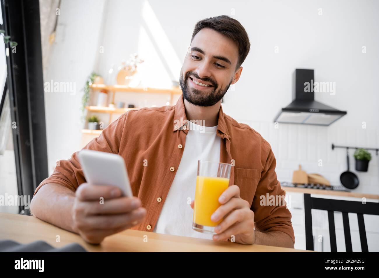hombre alegre y barbudo sosteniendo un vaso de zumo de naranja mientras utilizaba un smartphone, imagen de archivo Foto de stock