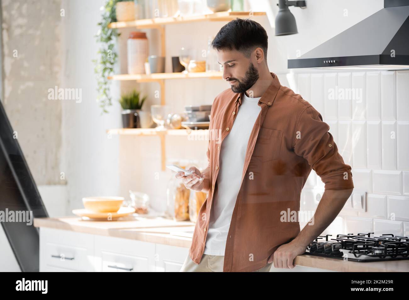 mensaje de hombre barbudo en el smartphone mientras se encuentra cerca de la cocina, imagen de archivo Foto de stock