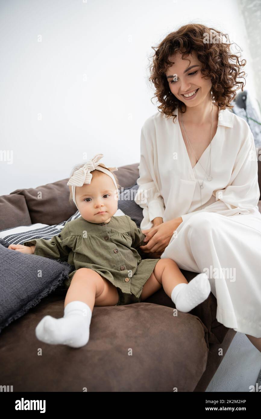 madre alegre en el vestido sonriendo cerca de la hija infantil en la diadema sentado en el sofá, imagen de archivo Foto de stock
