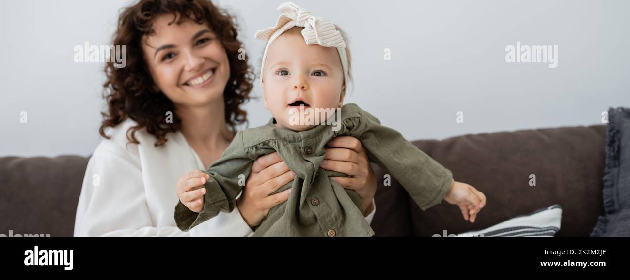 feliz madre con pelo rizado sonriendo mientras sostiene en brazos a la hija infantil en la diadema, banner, imagen de archivo Foto de stock