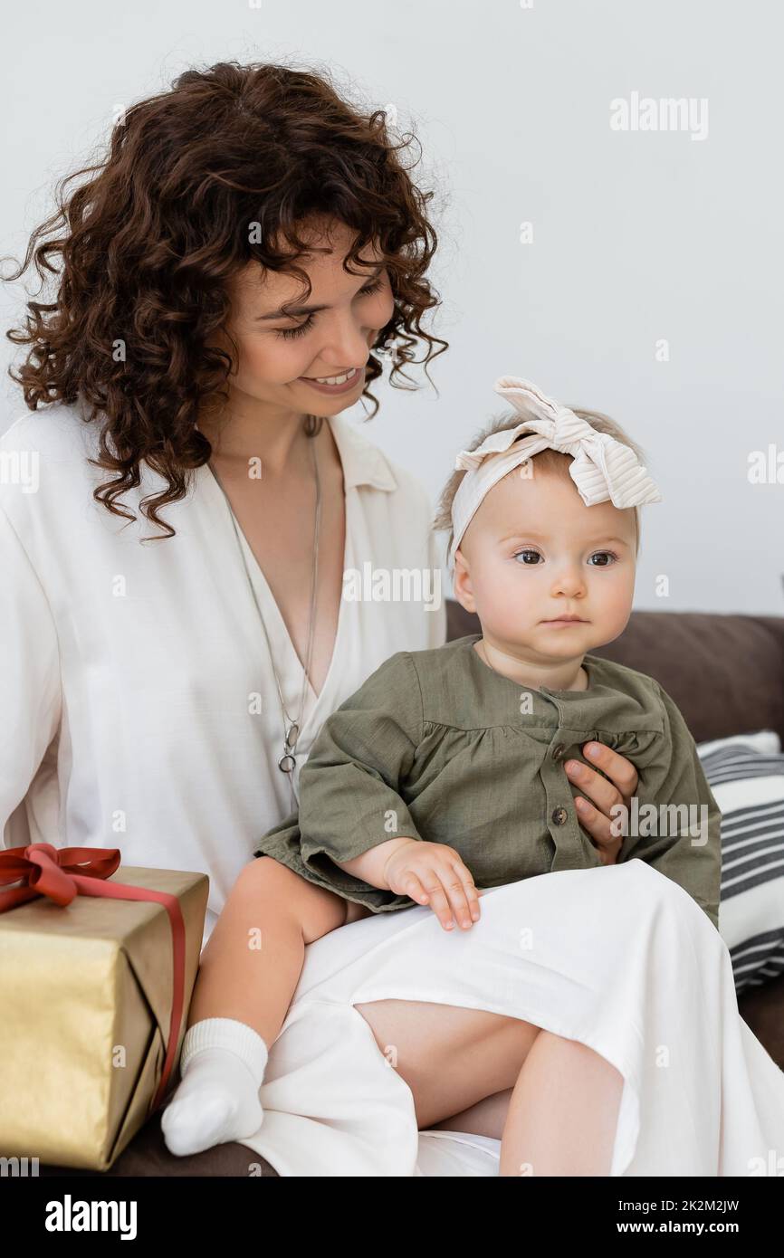 feliz y rizado madre en vestido sosteniendo caja de regalo envuelta cerca de la hija del bebé en diadema, imagen de archivo Foto de stock