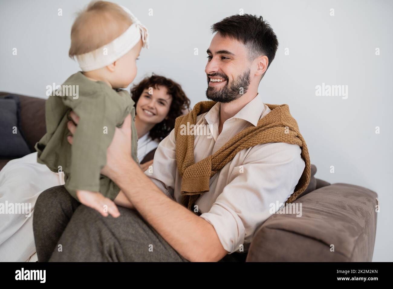 padre alegre sosteniendo a hija infantil cerca de feliz esposa borrosa en la sala de estar, imagen de archivo Foto de stock