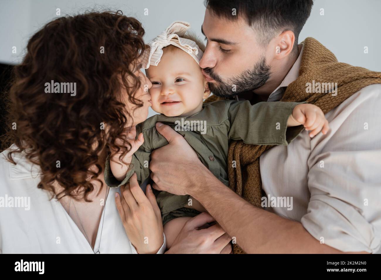 hombre barbudo y mujer rizada besando mejillas de la hija feliz bebé en diadema, imagen de archivo Foto de stock
