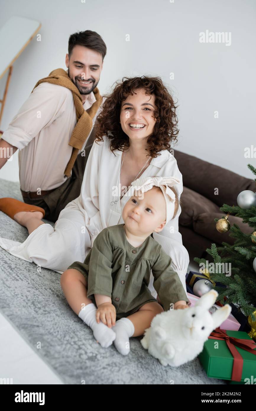 felices padres y lindo bebé sentado con juguete suave cerca de árbol de navidad decorado con regalos, imagen de archivo Foto de stock